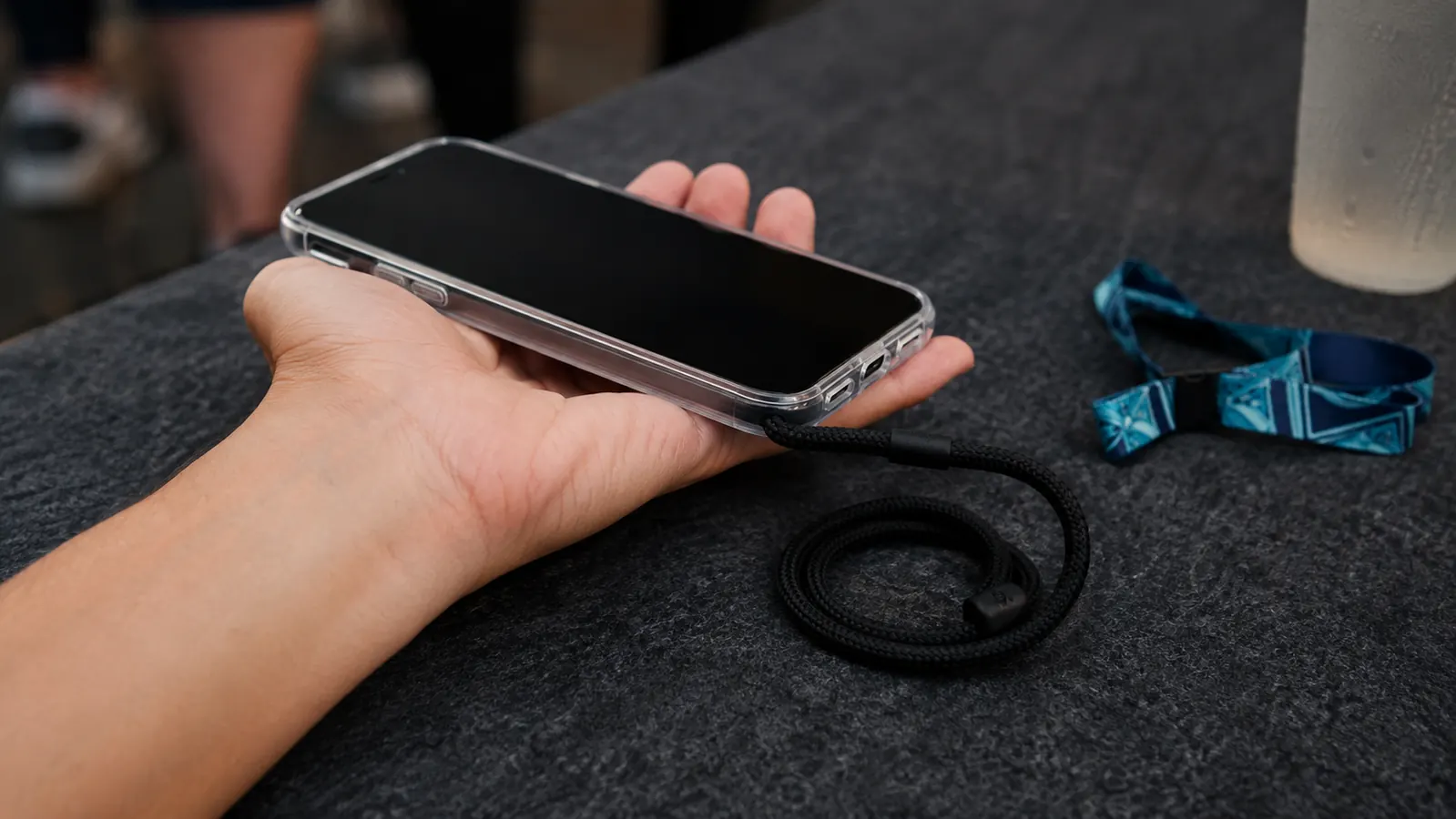 A phone resting in an open palm with the lanyard loop coiled beside it on a felt table top next to a folded festival wristband