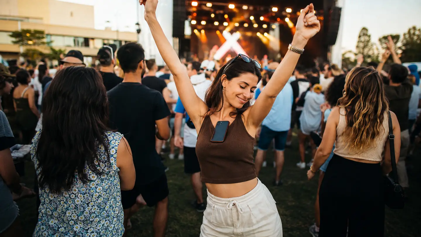 A person dancing at an outdoor festival, phone tucked against their chest, both hands raised free, late afternoon sunlight across the field