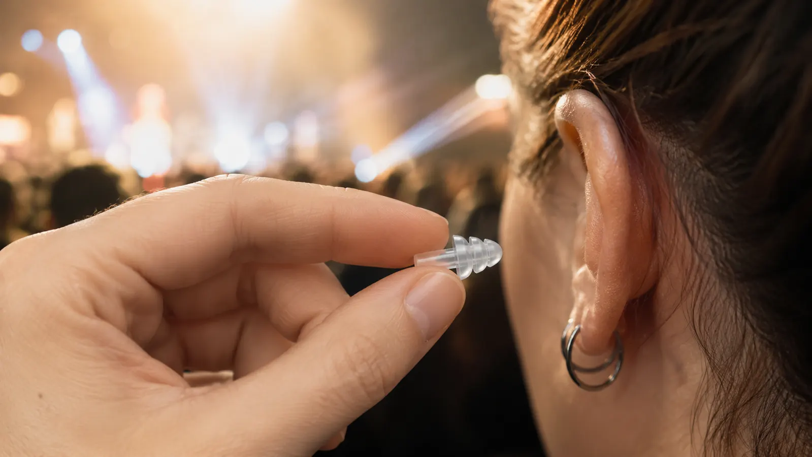 Close-up of a hand gently pressing a small high fidelity earplug into an ear in front of a softly lit concert crowd