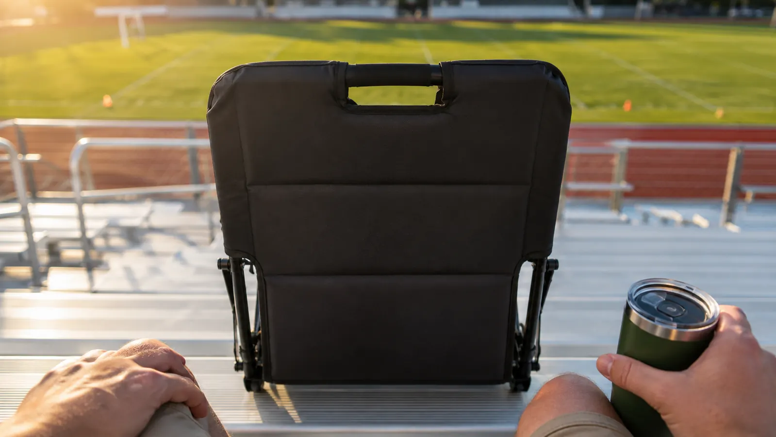 A padded folding stadium seat with a backrest and a carry handle sits on the metal bench of a bleacher row, seen from a seated height at a show.