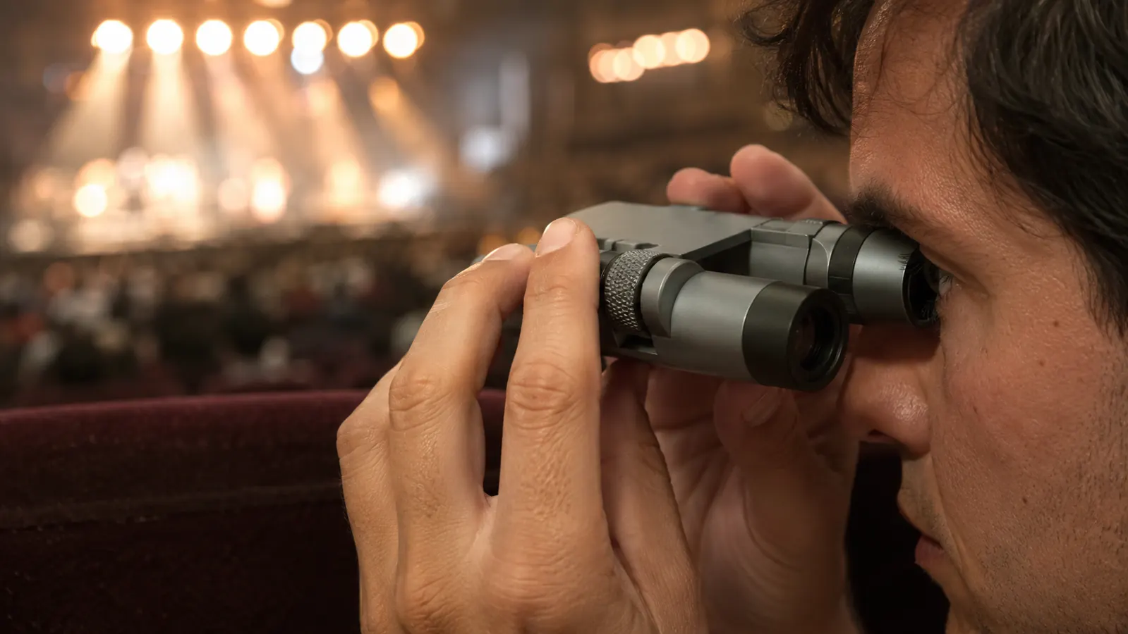 Close view of a compact binocular resting against an eye in two hands, blurred stage lights and crowd behind