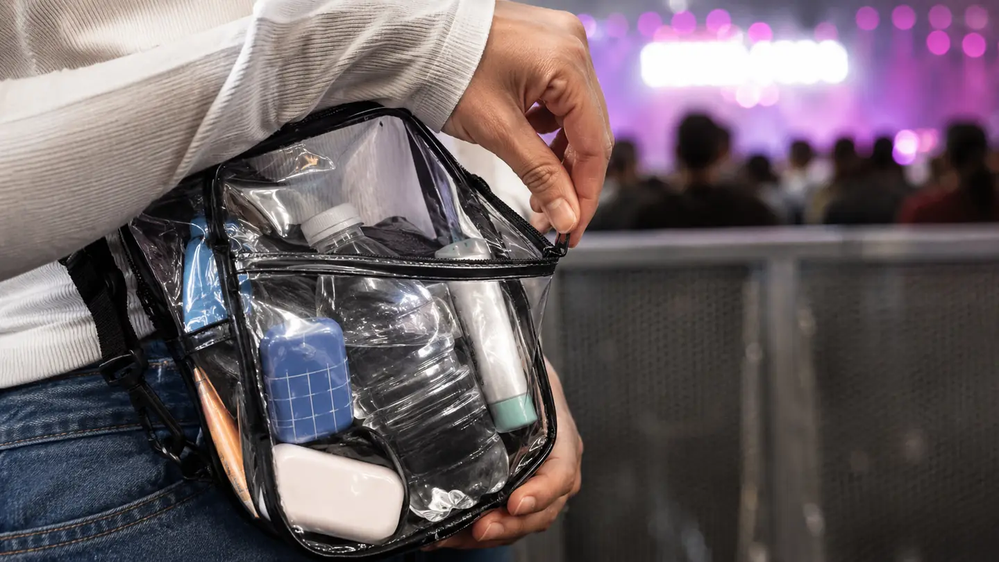 A hand closing the top zipper of the transparent bag that is packed full and snagging at the corner